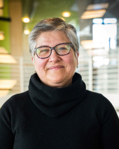 A close up portrait of Margaret E. Johnson wearing glasses & a black turtle nek, standing in front of an office-type background