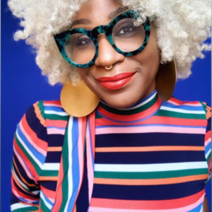 A close up portrait of Omisade Burney-Scott in a striped multicolor top, glasses, and standing in front of a vibrant blue backdrop